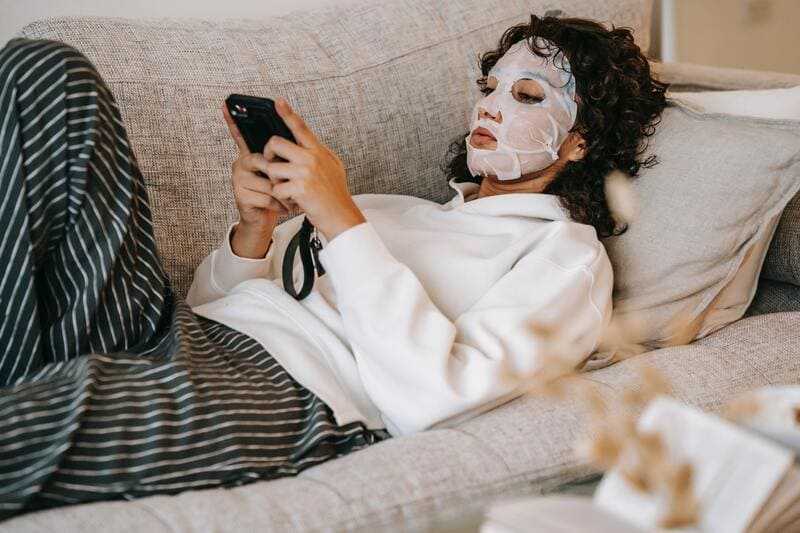 Woman lying on a couch with a facemask on -- she's researching alternative skincare treatments.